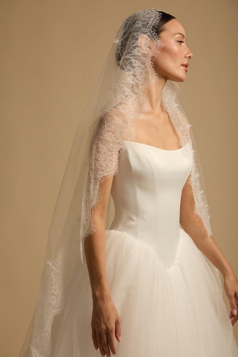 May include: A white wedding dress with a square neckline and a tulle skirt. The bride is wearing a delicate lace-trimmed veil. The veil is draped over the head and shoulders, creating a soft, romantic look. The dress is elegant and classic.