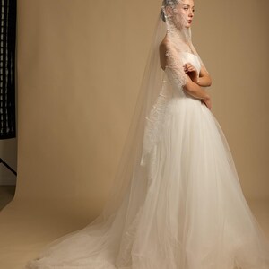 May include: A white wedding dress with a strapless bodice and a full, flowing skirt. The dress features delicate lace detailing and is paired with a long, sheer veil. The model is standing in a studio setting.