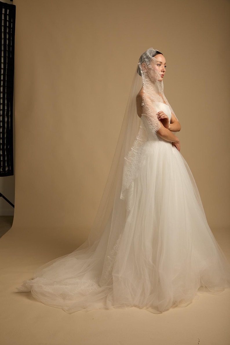 May include: A white wedding dress with a strapless bodice and a full, flowing skirt. The dress features delicate lace detailing and is paired with a long, sheer veil. The model is standing in a studio setting.
