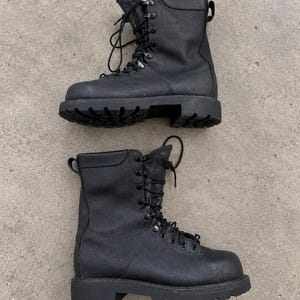 May include: A pair of black leather lace-up boots featuring a high-top design, a rounded toe, and a thick, lugged rubber sole. The boots include metal eyelets and hooks for the laces, a padded collar, and a pull tab at the back of the ankle.
