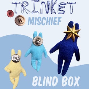 May include: A light blue background features the words "TRINKET MISCHIEF BLIND BOX". Three felt figures in yellow, light blue, and dark blue are shown. Each figure has a button for a face. A gold star is attached to the dark blue figure.