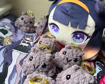 May include: A group of purple crocheted plushies with gold halo accents. The plushies have black eyes and a small smile. The plushies are sitting on a black and white patterned surface.