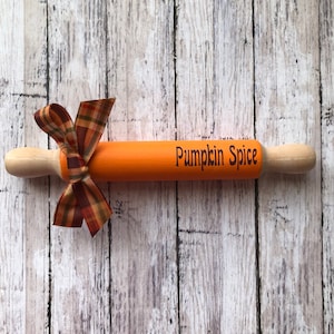 May include: Orange wooden rolling pin with a brown and orange plaid ribbon tied around it. The rolling pin is decorated with the words "Pumpkin Spice" in black.