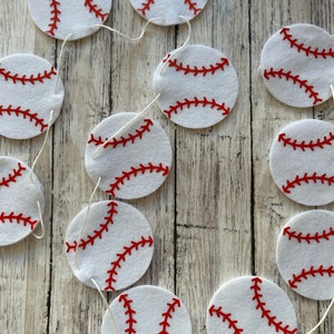 May include: A white felt baseball garland with red stitching. The garland is strung together with white twine and has 10 baseballs.