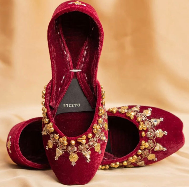 May include: A pair of maroon velvet shoes with gold embroidery and embellishments. The shoes have a pointed toe and a low heel. The brand name "DAZZLE" is visible on the inside of one shoe.