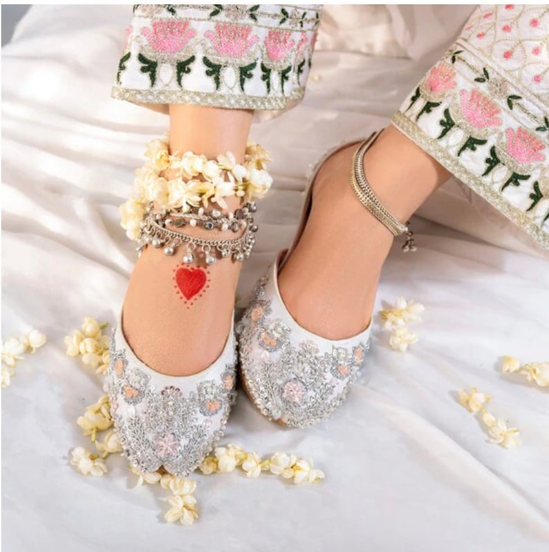 K&ouml;nnte beinhalten: Wei&szlig;e bestickte flache Schuhe mit silbernen Verzierungen. Die Schuhe haben eine spitze Zehenpartie und sind mit Blumenmustern verziert. Die Schuhe werden von einer Person mit silbernen Fu&szlig;kettchen und einem roten Herz getragen, das auf ihren Kn&ouml;chel gezeichnet ist.