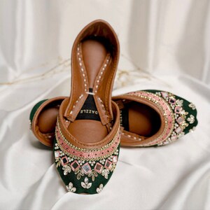 Puede incluir: Un par de zapatos de terciopelo verde con bordados en rosa y dorado y adornos de espejo. Los zapatos tienen una plantilla de cuero marr&oacute;n con la palabra "DAZZLE" impresa en ella.