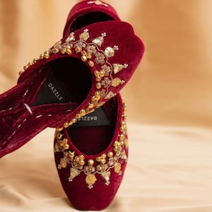 May include: A pair of maroon velvet slippers with gold and silver embellishments. The slippers have a pointed toe and a low heel. The brand name "DAZZLE" is visible on the insole.