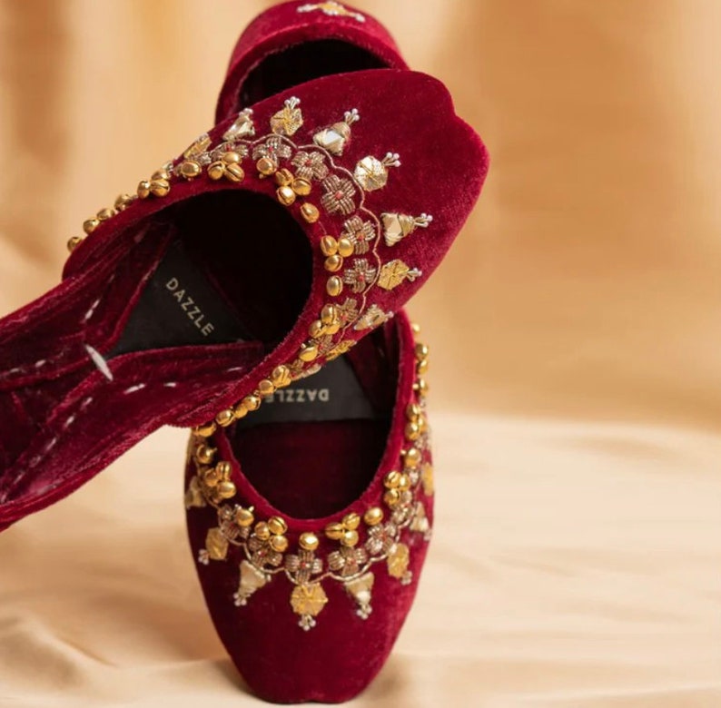 May include: A pair of maroon velvet slippers with gold and silver embellishments. The slippers have a pointed toe and a low heel. The brand name "DAZZLE" is visible on the insole.