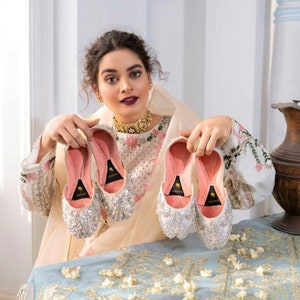 K&ouml;nnte beinhalten: Ein Paar wei&szlig;e, verzierte Hochzeitsschuhe mit rosa Futter. Die Schuhe sind mit Pailletten verziert und haben eine spitze Zehenpartie. Die Schuhe werden von einer Person in einem wei&szlig;en, bestickten Kleid gehalten.
