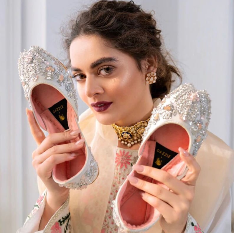 K&ouml;nnte beinhalten: Ein Paar wei&szlig;e, flache Schuhe mit Stickereien in Silber und Wei&szlig;. Die Schuhe haben ein rosafarbenes Futter und das Wort "DAZZLE" ist auf der Innenseite jedes Schuhs aufgedruckt.