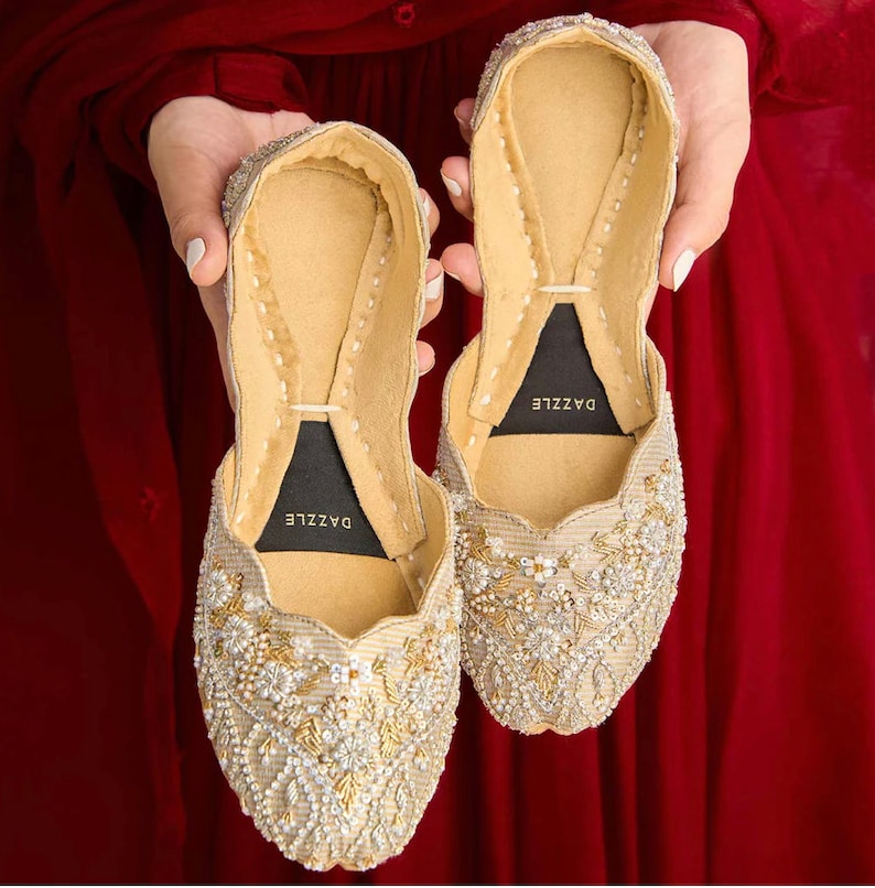 Puede incluir: Un par de zapatos planos dorados adornados con cuentas con una etiqueta negra que dice "DAZZLE" en el interior de cada zapato.