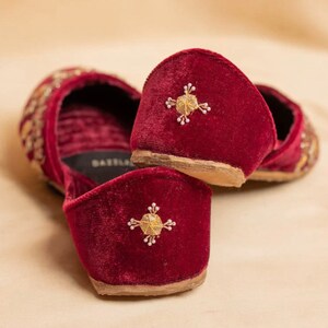 May include: A pair of burgundy velvet slippers with gold embroidery. The slippers have a pointed toe and a low heel. The embroidery features a geometric design with a central star.