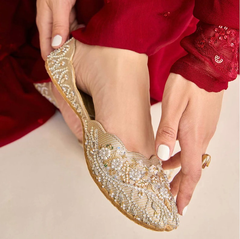 Puede incluir: Un par de zapatos planos bordados en oro con adornos de perlas y cristales. Los zapatos est&aacute;n siendo puestos por una persona que lleva un atuendo rojo.