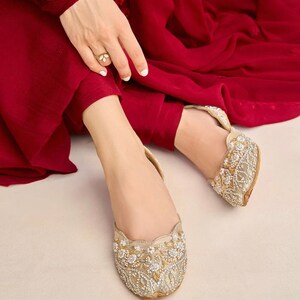 Puede incluir: Un par de zapatos planos dorados adornados con cuentas blancas y dise&ntilde;os florales. Los zapatos son usados por una persona que lleva un atuendo rojo.