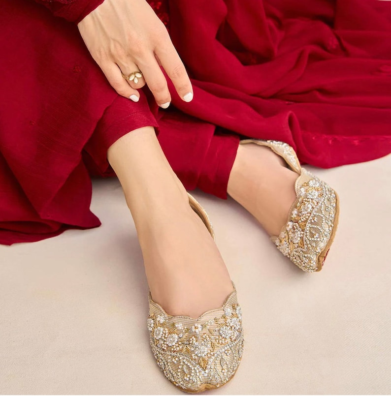 Puede incluir: Un par de zapatos planos dorados adornados con cuentas blancas y dise&ntilde;os florales. Los zapatos son usados por una persona que lleva un atuendo rojo.