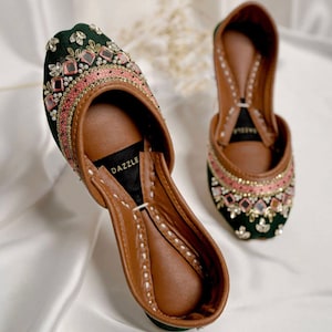 Puede incluir: Un par de zapatos de terciopelo verde con bordados intrincados y adornos de espejo. Los zapatos tienen un ribete rosa y una plantilla de cuero marr&oacute;n con la palabra "DAZZLE" impresa.