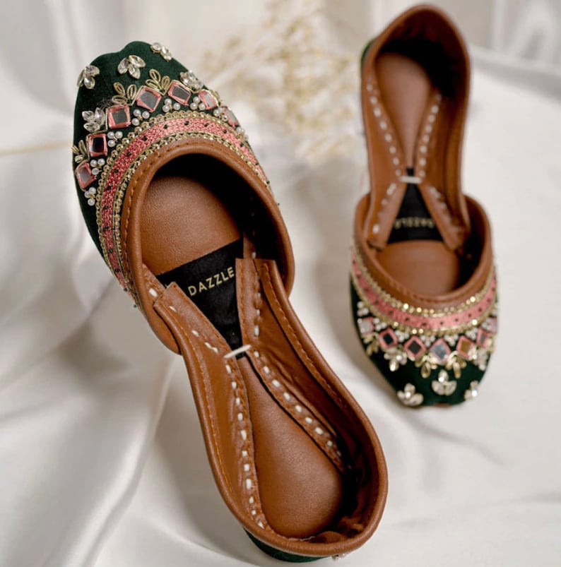 Puede incluir: Un par de zapatos de terciopelo verde con bordados intrincados y adornos de espejo. Los zapatos tienen un ribete rosa y una plantilla de cuero marr&oacute;n con la palabra "DAZZLE" impresa.