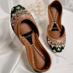 Puede incluir: Un par de zapatos de terciopelo verde con bordados rosa y dorados y adornos de espejo. Los zapatos tienen una plantilla de cuero marr&oacute;n y una punta puntiaguda. El nombre de la marca "DAZZLE" es visible en la plantilla.