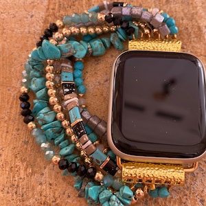 May include: A gold-toned smartwatch with a black screen is paired with a stack of beaded bracelets. The bracelets feature turquoise, black, gold, and neutral-toned beads. The watch band has a textured design.
