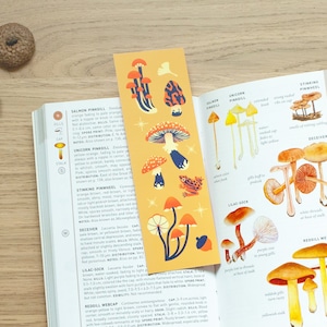 Könnte beinhalten: Ein gelbes Lesezeichen mit einer bunten Illustration von Pilzen. Das Lesezeichen liegt auf einem geöffneten Buch über Pilze. Das Buch enthält Illustrationen verschiedener Pilzarten mit ihren Namen und Beschreibungen.