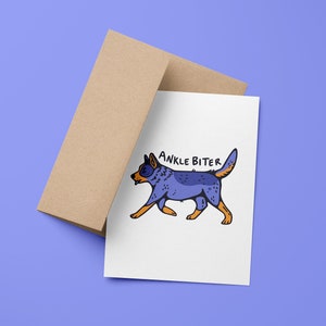 Puede incluir: Una tarjeta blanca con una ilustraci&oacute;n de perro de dibujos animados azul y naranja y el texto "ANKLE BITER" sobre un fondo morado claro. La tarjeta est&aacute; encima de un sobre marr&oacute;n.