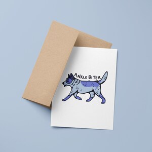 Puede incluir: Una tarjeta blanca con una ilustraci&oacute;n de perro de dibujos animados azul y gris y el texto "ANKLE BITER" en negro. La tarjeta est&aacute; encima de un sobre marr&oacute;n.