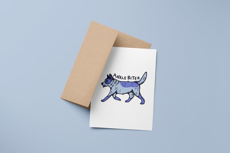 Puede incluir: Una tarjeta blanca con una ilustraci&oacute;n de perro de dibujos animados azul y gris y el texto "ANKLE BITER" en negro. La tarjeta est&aacute; encima de un sobre marr&oacute;n.