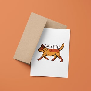 Puede incluir: Una tarjeta blanca con una ilustraci&oacute;n de dibujos animados de un perro rojo y naranja con el texto "ANKLE BITER" encima. La tarjeta est&aacute; encima de un sobre marr&oacute;n.