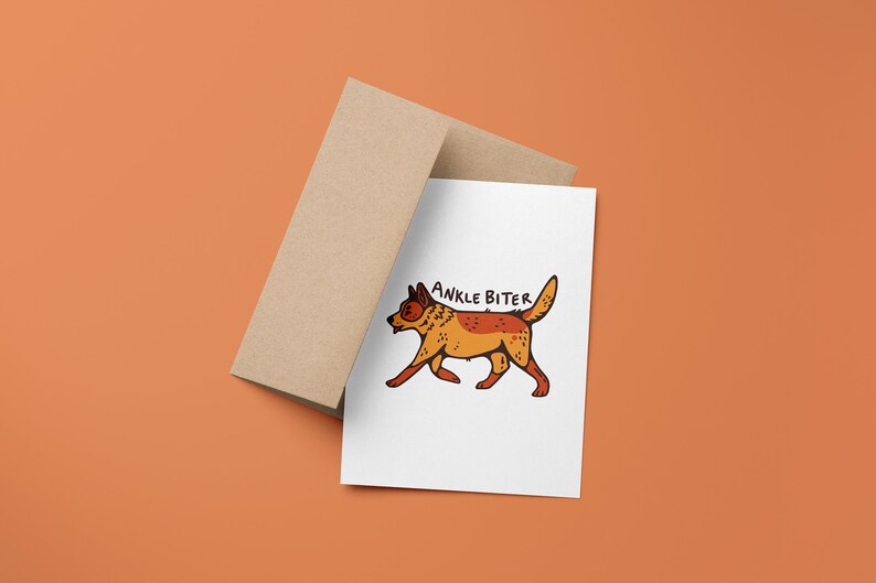 Puede incluir: Una tarjeta blanca con una ilustraci&oacute;n de dibujos animados de un perro rojo y naranja con el texto "ANKLE BITER" encima. La tarjeta est&aacute; encima de un sobre marr&oacute;n.