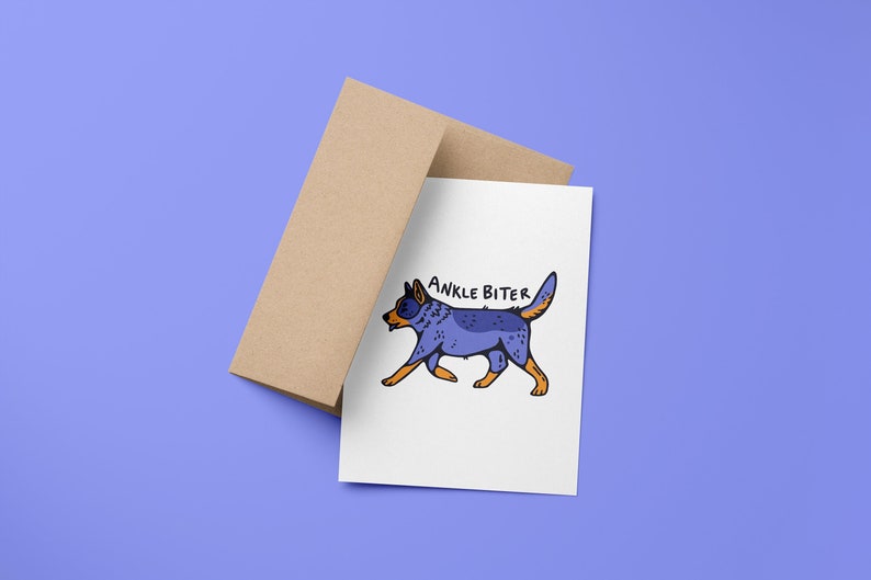 Puede incluir: Una tarjeta blanca con una ilustraci&oacute;n de perro de dibujos animados azul y naranja y el texto "ANKLE BITER" sobre un fondo morado claro. La tarjeta est&aacute; encima de un sobre marr&oacute;n.