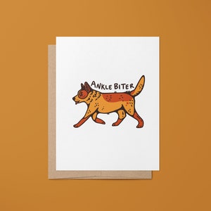 Puede incluir: Una tarjeta blanca con un sobre marr&oacute;n. La tarjeta presenta una ilustraci&oacute;n de dibujos animados de un perro rojo y naranja con el texto "ANKLE BITER" encima.