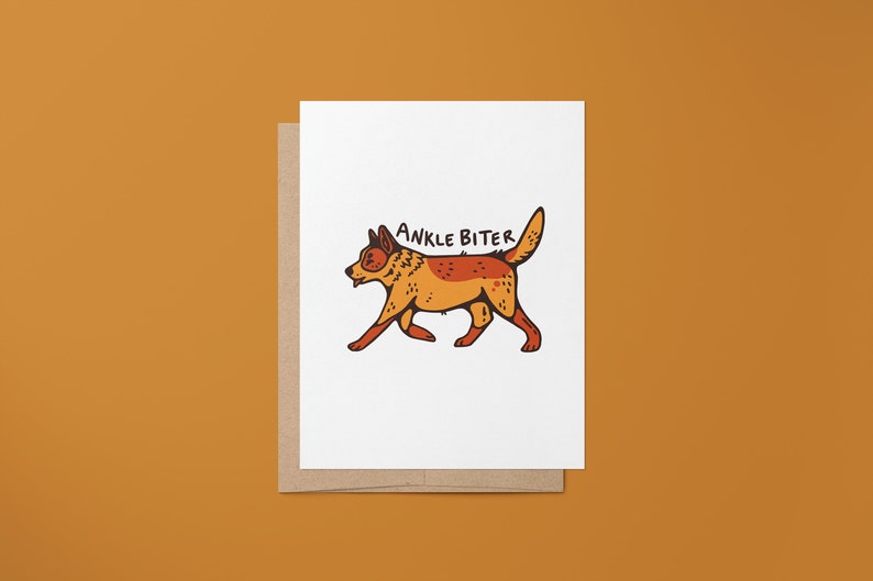 Puede incluir: Una tarjeta blanca con un sobre marr&oacute;n. La tarjeta presenta una ilustraci&oacute;n de dibujos animados de un perro rojo y naranja con el texto "ANKLE BITER" encima.
