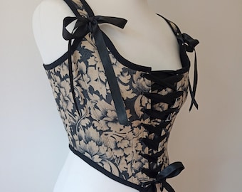 May include: A black and gold corset with a floral pattern and black ribbon ties. The corset has a lace-up front and a fitted silhouette.