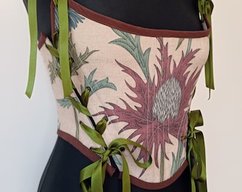 May include: A beige corset with a floral print of green and red thistles. The corset has brown trim and green satin ribbon laces.