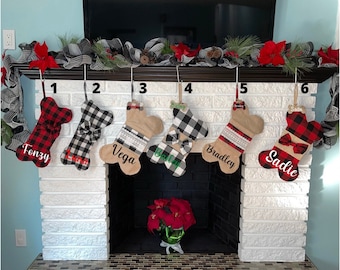 May include: Six personalized dog bone-shaped Christmas stockings hanging on a white brick fireplace mantel. The stockings are made of burlap and red and black buffalo plaid fabric. The stockings are decorated with bows and have the names Fonzy, Luna, Vega, Bailey, Bradley, and Sadie embroidered on them.