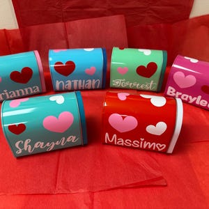 Op de afbeelding: Zes kleine, kleurrijke metalen brievenbussen met hartjesontwerpen en gepersonaliseerde namen: Brianna, Nathan, Forrest, Braylee, Shayna en Massimo.