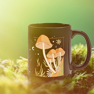 May include: Black ceramic mug featuring a whimsical illustration of mushrooms with a celestial design. The artwork includes tan and orange mushrooms, stars, and a crescent moon. The mug has a black handle and is set against a mossy background.