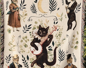May include: A woven tapestry featuring a whimsical scene of cats playing music. The tapestry is cream-colored with green foliage and crescent moons. One cat plays a banjo, another plays a violin, and a third cat wears a top hat and holds a cane.
