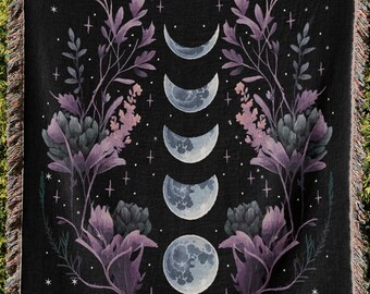 May include: A black woven throw blanket featuring a central vertical sequence of five moon phases, flanked by symmetrical illustrations of purple and grey botanical floral arrangements and scattered white stars, finished with a fringed edge.