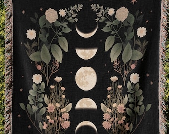 May include: A black woven throw blanket featuring a central vertical column of five moon phases. The design is flanked by botanical illustrations of flowers and leaves in muted tones, accented with small four-pointed stars throughout the fabric.