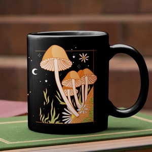 May include: Black ceramic mug featuring a whimsical illustration of orange and tan mushrooms against a starry night sky with a crescent moon and stars. The mug is placed on a stack of books with green and brown covers.