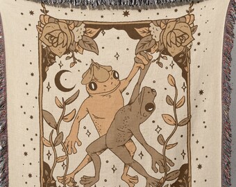 May include: A woven tapestry featuring a tarot card design with two frogs dancing. The card is titled "THE LOVERS" and is surrounded by stars, roses, and leaves. The tapestry is beige with brown accents.