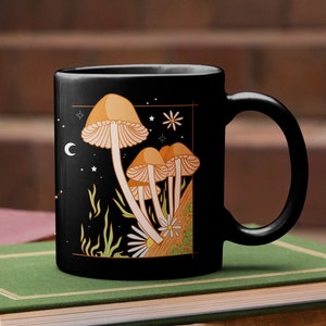 May include: Black ceramic mug featuring a whimsical illustration of orange mushrooms with white gills, a daisy, and stars against a night sky. The mug has a curved handle and is likely used for hot beverages.