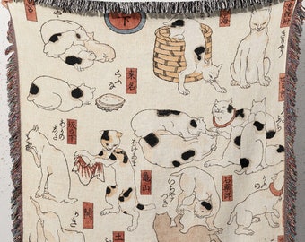 May include: A woven tapestry throw blanket featuring a vintage Japanese illustration of cats in various poses. The design is in black, white, and red on a cream background. The Japanese text on the blanket reads "猫の図" which translates to "Cat Illustration".