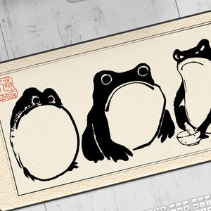 May include: A black and white mousepad with a design of three frogs. The frogs are in different poses and have a simple, stylized design. The background is a light beige with a pattern of waves.