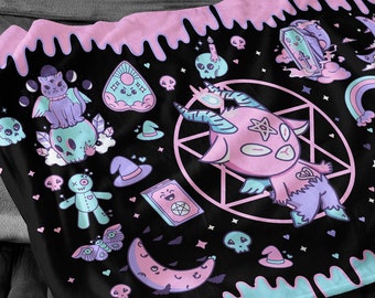 May include: A black and pink throw blanket with a dripping paint design. The blanket features a variety of cute, spooky, and gothic illustrations, including a pink demon, a skull, a witch's hat, and a crescent moon.