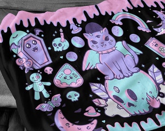 May include: Black blanket with a pastel goth design featuring a cat, skull, coffin, and other mystical elements. The blanket has a pink and blue border and is displayed on a bed with gray bedding.
