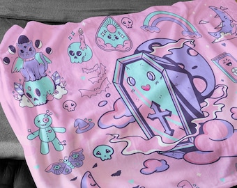 May include: Pink throw blanket with a cute, spooky design featuring a coffin, a cat, a voodoo doll, a butterfly, and a crescent moon. The design is on a pink background with white and purple accents.