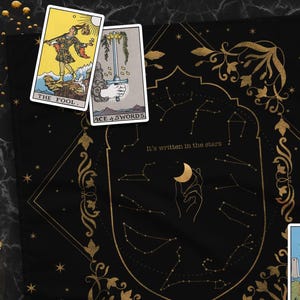 May include: A black tarot cloth with gold constellation designs and the text "It's written in the stars." Several tarot cards are scattered across the cloth, including "The Fool," "Ace of Swords," and "The Moon." A purple crystal is also present.